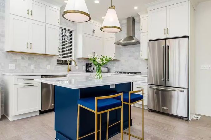 Bright modern kitchen with white shaker cabinets and marble countertop island in navy, two gold-framed blue bar stools, stainless steel French-door refrigerator, brass faucet, pendant lights, and light hardwood floors.