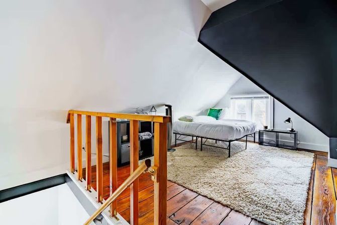 Cozy attic loft bedroom with sloped white ceiling, dark accent wall, wooden plank floors, large shag rug and minimalist bed by a dormer window