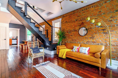 Cozy urban loft living room with exposed red brick wall, tan leather sofa with colorful pillows and yellow throw, rattan chair, dark open staircase, glossy hardwood floors, brass arc floor lamp, round wall mirror and potted plant.