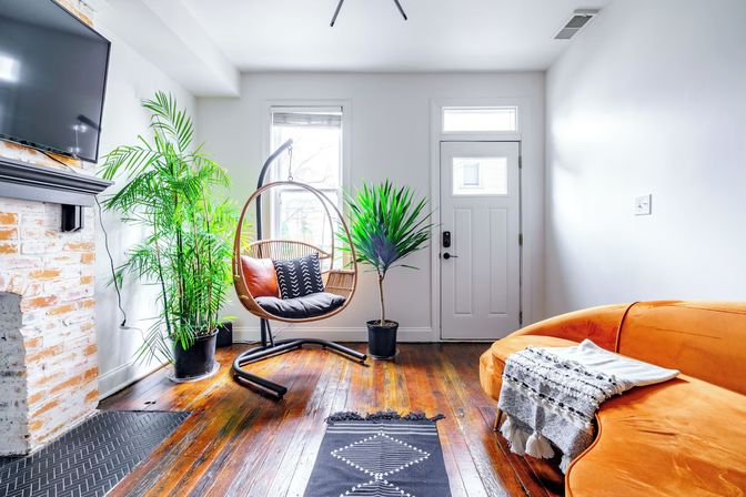 Sunlit modern living room with hardwood floors, orange velvet sofa, hanging rattan egg chair with patterned cushions, potted tropical plants, white entry door and whitewashed brick fireplace with wall-mounted TV.