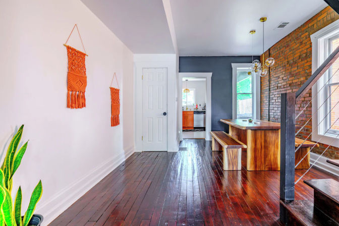 Sunlit urban townhouse dining nook with exposed brick wall, live-edge wooden table and benches, globe pendant lights, polished dark hardwood floors, orange macramé wall hangings and view into a bright white kitchen.