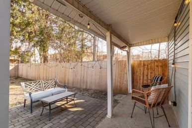 Cozy suburban backyard patio with covered porch, string lights, paver floor, metal bench and coffee table, two wicker chairs with cushions beside a wooden privacy fence and trees