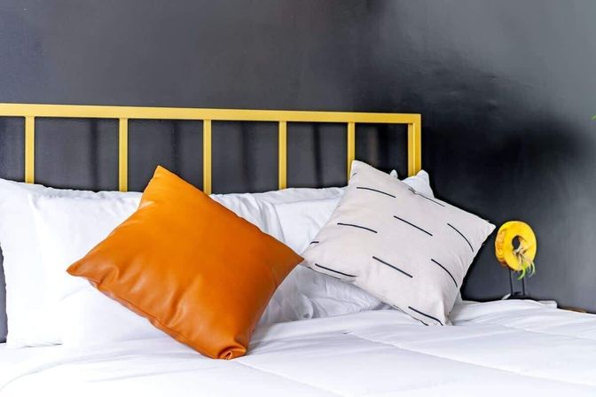 Modern bedroom interior with yellow metal headboard against a dark gray wall, crisp white bedding and a pop of color from an orange leather throw pillow and a striped accent cushion.