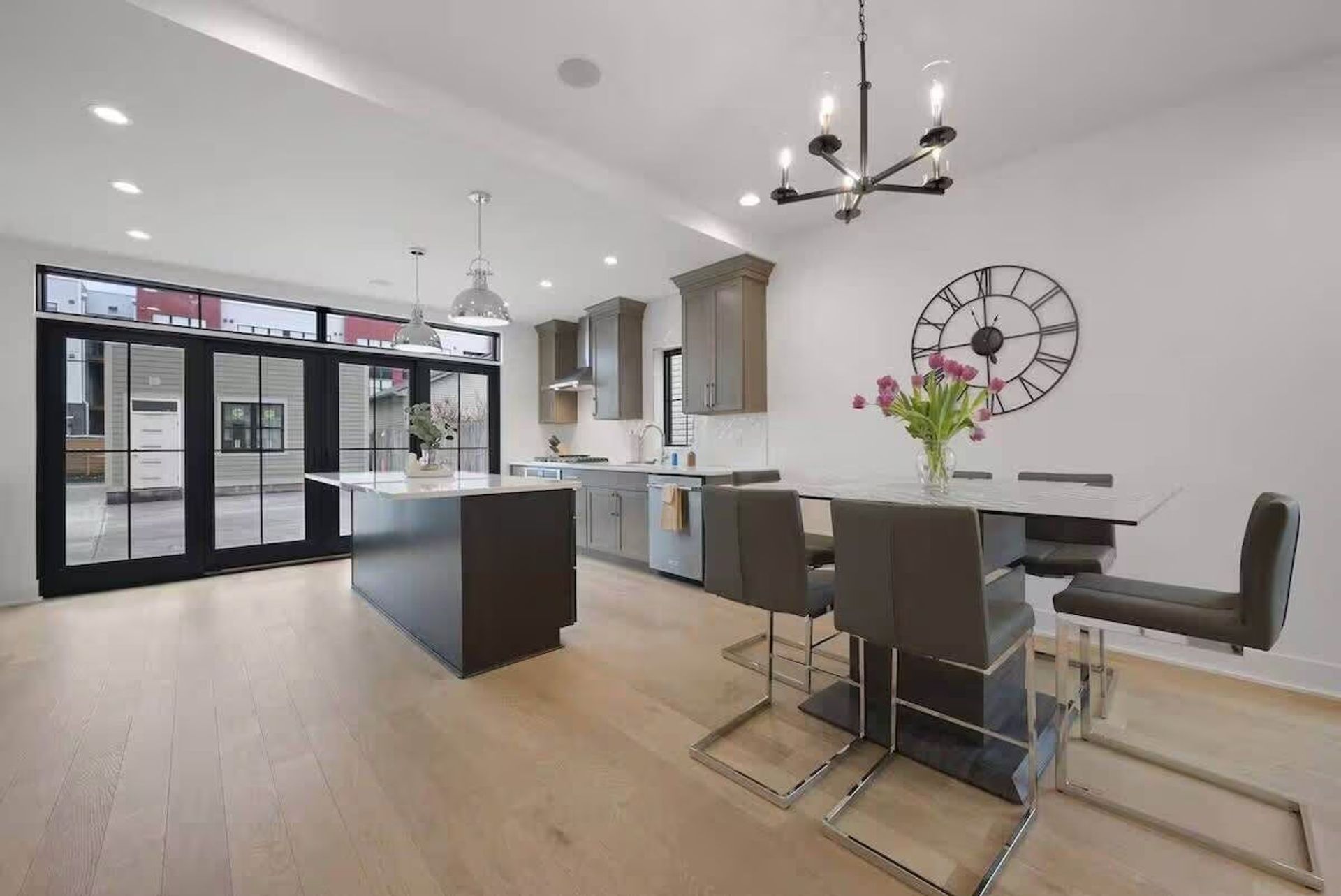 Bright modern open-plan kitchen and dining area with a central island, gray cabinets, black-framed floor-to-ceiling sliding doors, light hardwood floors, chrome pendant and chandelier lighting, wall clock, and a dining table with gray leather chairs and pink tulips.