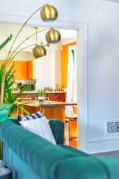 Open-concept living room and kitchen with emerald green velvet sofa in foreground, gold arc floor lamp, indoor plants, wooden island and bright orange cabinets in a sunny modern interior