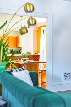 Open-concept living room and kitchen with emerald green velvet sofa in foreground, gold arc floor lamp, indoor plants, wooden island and bright orange cabinets in a sunny modern interior