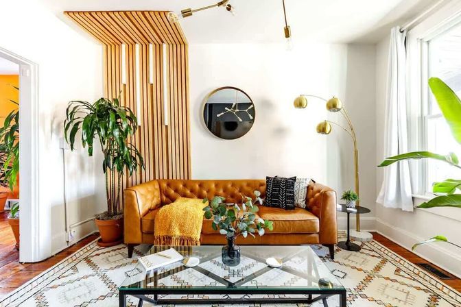 Bright modern living room featuring a tan tufted leather sofa, vertical wooden slat accent wall, brass arc floor lamp, indoor plants, glass coffee table and geometric area rug by a sunlit window.