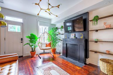 Sunlit, plant-filled modern living room with hardwood floors, a rattan hanging chair, black fireplace topped by a wall-mounted TV and a front entry door.