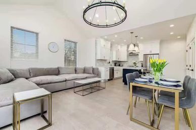 Bright open-concept living room and kitchen with vaulted ceiling, large gray sectional sofa, minimalist metal coffee table, gold-accent dining table set with yellow tulips, white cabinets, stainless steel appliances, and pendant lighting.