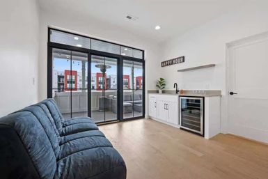 Bright modern apartment living room — plush blue sofa, hardwood floors, wet bar with wine fridge and 'Happy Hour' sign, sliding glass doors to balcony with urban city view