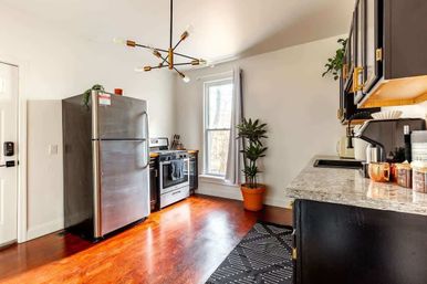 Sunlit modern kitchen with stainless steel refrigerator and stove, black cabinets, marble countertop, warm hardwood floors, potted plant, and mid-century pendant light.
