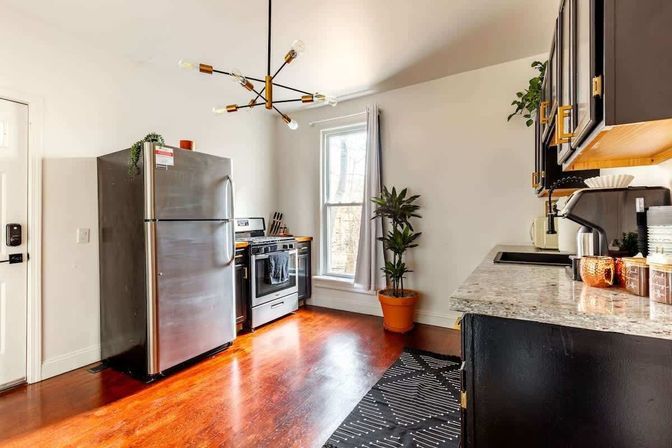 Sunlit modern kitchen with stainless steel refrigerator and stove, black cabinets, marble countertop, warm hardwood floors, potted plant, and mid-century pendant light.