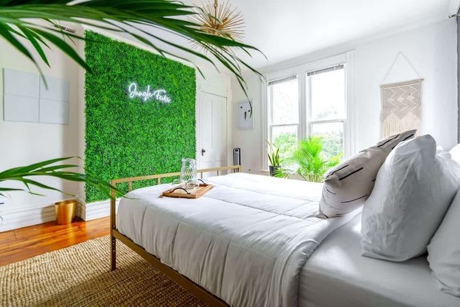 Bright sunlit bedroom with white bedding and gold metal bed frame, vibrant green living wall with neon sign, indoor potted plants, hardwood floor and boho accents