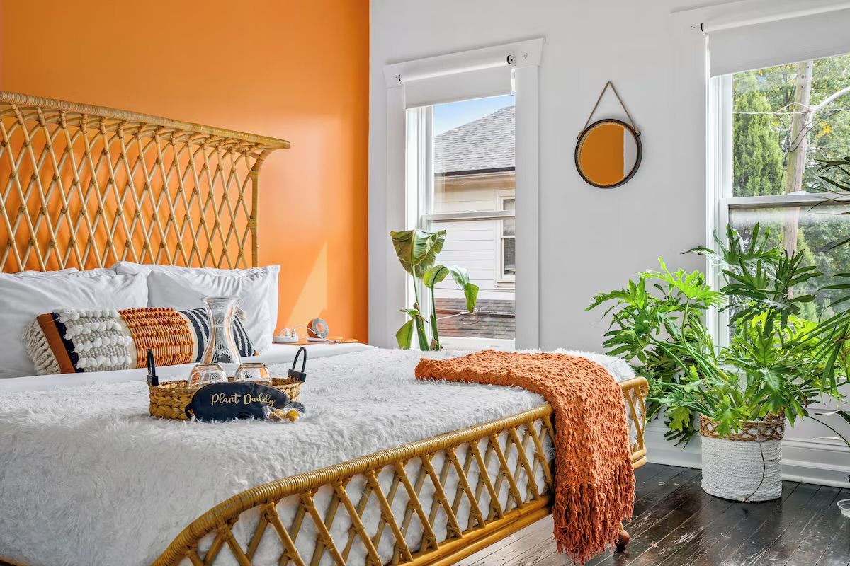 Sunlit boho bedroom with a bright orange accent wall, woven rattan bedframe, white fluffy bedding, textured accent pillows and orange throw, round wall mirror and lush potted plants by two windows.