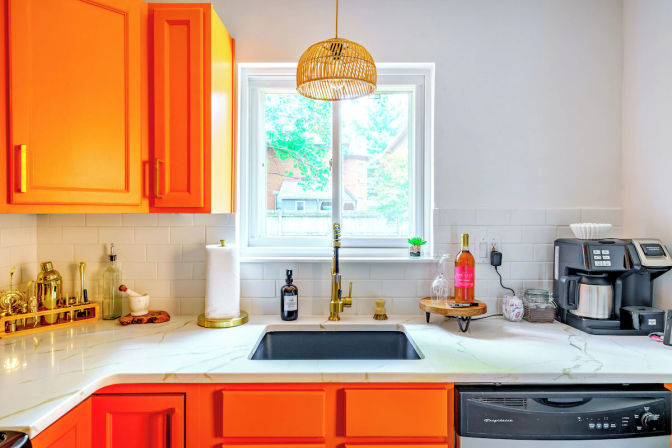 Vibrant modern kitchen with bold orange cabinets, marble-look countertop, black undermount sink under a window, brass faucet and rattan pendant light, white subway tile backsplash, coffee maker and wine bottle on the counter.