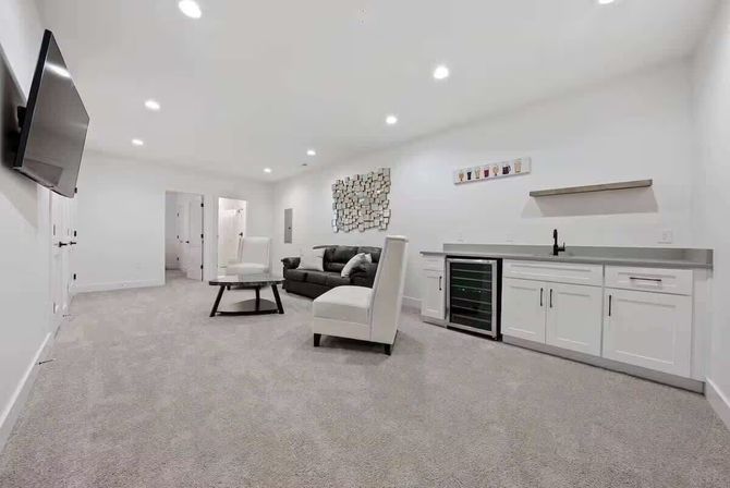 Cozy modern finished basement lounge with gray carpet, white walls, recessed lighting, wall-mounted TV, dark sofa and white chairs around a round coffee table, and a built-in wet bar with wine fridge and stainless sink.