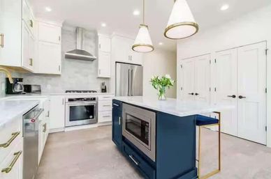 Bright modern white kitchen with a navy island and built-in microwave, marble countertops, stainless steel fridge and range, brass hardware, pendant lights and a vase of flowers