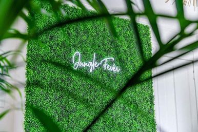 Indoor green boxwood wall with neon sign reading "Jungle Fever", framed by blurred tropical palm leaves for a modern indoor jungle decor backdrop