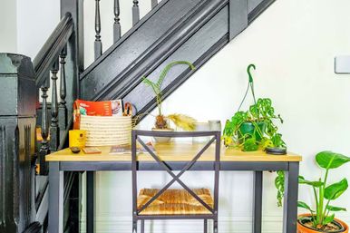 Cozy home-office nook under a black staircase banister featuring a wooden desk with a woven-seat chair, hanging and potted plants, and a woven basket of magazines and mail.