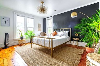 Airy sunlit modern boho bedroom with a gold metal bed frame, black accent wall, oversized potted plants, white bedding and a jute rug on polished hardwood floors