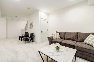 Bright, cozy modern finished basement living room with a gray sectional sofa and patterned pillows, marble-top coffee table with a small vase of flowers, two dark accent chairs by a staircase, and white walls.