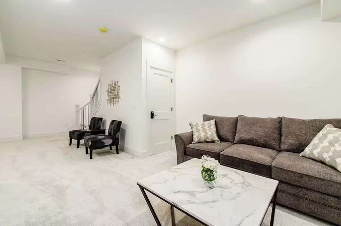 Bright, cozy modern finished basement living room with a gray sectional sofa and patterned pillows, marble-top coffee table with a small vase of flowers, two dark accent chairs by a staircase, and white walls.