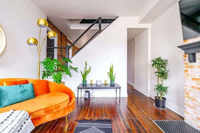 Bright modern living room interior with a curved orange velvet sofa and teal pillow, brass arc floor lamp, multiple potted plants, black metal staircase, exposed brick fireplace, and polished hardwood floors.