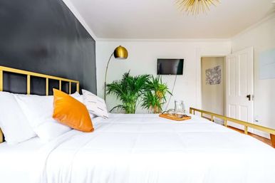 Bright modern apartment bedroom with white bedding and gold metal bed frame, orange accent pillow, black accent wall, indoor palm plants, wall-mounted TV and gold arc floor lamp.