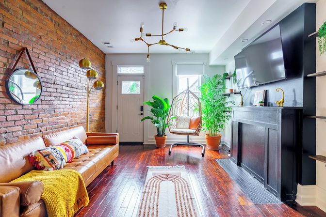 Sunny urban living room with exposed brick wall, caramel leather sofa with colorful throw, brass floor lamp and modern chandelier, hanging rattan egg chair amid potted plants on dark hardwood floors, TV mounted above a black mantel