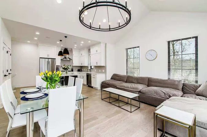 Bright open-concept living room and kitchen with light wood floors, gray sectional sofa, glass dining table with white chairs and yellow tulips, and stainless appliances.