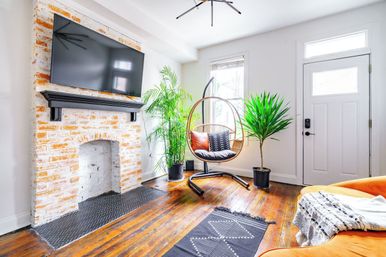 Sunlit modern living room with whitewashed brick fireplace and wall-mounted TV, hanging rattan egg chair with cushions, hardwood floors, potted indoor plants, and a bright orange sofa by a white front door.