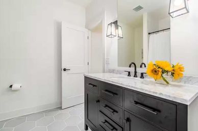 Bright modern bathroom with a black vanity and marble countertop, black faucet and hardware, gray hexagon floor tiles, pendant lights, and sunflowers in a glass vase.
