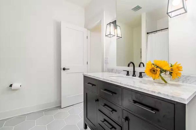 Bright modern bathroom with a black vanity and marble countertop, black faucet and hardware, gray hexagon floor tiles, pendant lights, and sunflowers in a glass vase.