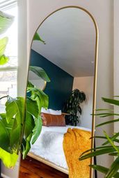 Gold arched floor mirror reflecting a cozy urban bedroom with a teal accent wall, lush houseplants, white faux-fur bedding, tan leather bolster pillow and a mustard orange throw on warm wood floors.