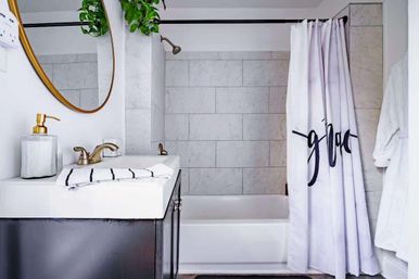 Bright modern bathroom with marble-look tiled tub/shower, white shower curtain with black script, dark vanity with white sink and gold faucet, round gold mirror and hanging green plant