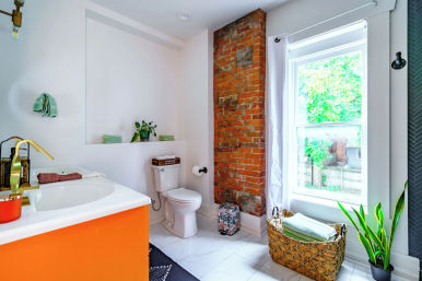 Sunlit modern bathroom with exposed red brick column, bold orange vanity and gold faucet, white marble floor, large window overlooking a green yard, woven towel basket and potted plants.