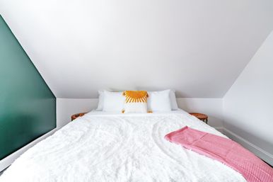 Cozy bright loft bedroom with sloped white ceiling, plush white bedspread, yellow sun-shaped accent pillow, pink throw and green accent wall