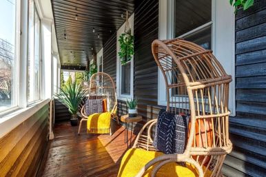 Sunlit enclosed porch with rattan egg chairs, bright yellow throws and patterned cushions, hardwood floor, hanging plants, small round side table and large windows.