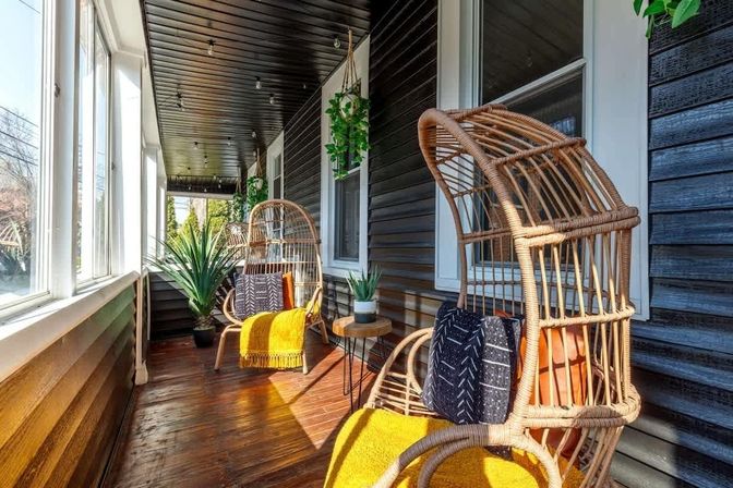 Sunlit enclosed porch with rattan egg chairs, bright yellow throws and patterned cushions, hardwood floor, hanging plants, small round side table and large windows.