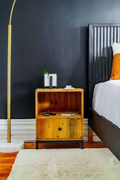 Contemporary bedroom corner in an urban apartment: black accent wall, wooden nightstand with small succulent planter, remote and notebook, gold floor lamp, white bed with orange pillow and plush rug.
