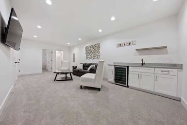 Bright, modern finished basement entertainment room — cozy seating, coffee table, wall-mounted TV, wet bar with wine fridge and white cabinets, gray carpet, recessed lighting.