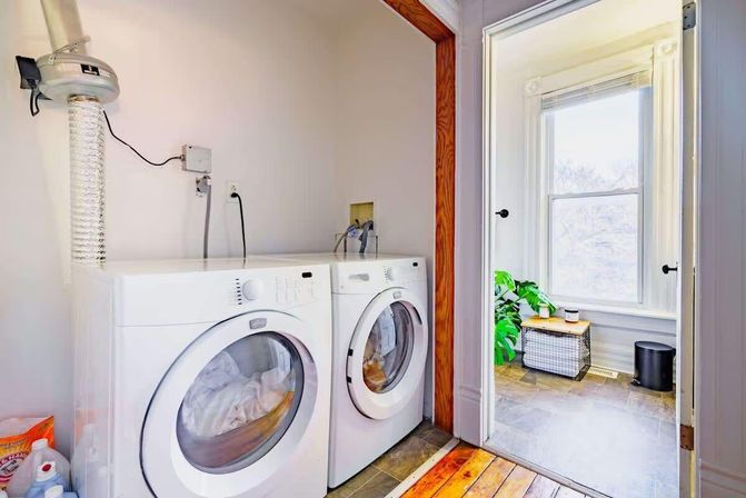 Cheerful home laundry nook with white front-loading washer and dryer, vent hose and detergent, opening onto a sunlit bay-windowed room with a potted plant on a wicker basket and mixed wood-and-tile flooring.