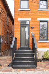 Charming red-brick urban rowhouse entrance with black wooden steps and railing leading to a glass front door, gray-trimmed windows, iron side gate and brick sidewalk.