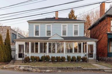 Charming two-story light blue house with an enclosed glass front porch, symmetrical upper windows, low shrubs, stone curb and overhead utility lines on a quiet residential street.