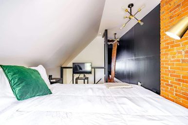 Urban loft bedroom with sloped ceiling, exposed brick and black accent wall, white bedding with a green pillow, wall-mounted TV and modern brass light fixtures.