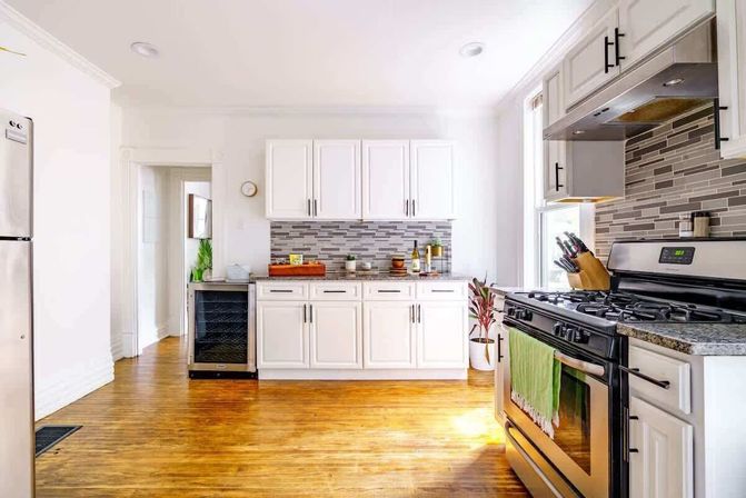 Sunny modern kitchen with white cabinets, gray mosaic tile backsplash, stainless steel fridge and gas range, small wine fridge, hardwood floors and potted plant