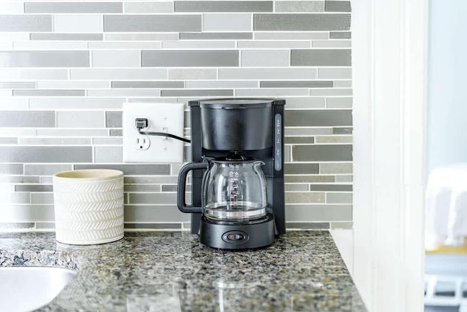 Black drip coffee maker with glass carafe on a speckled granite kitchen counter, set against a gray linear tile backsplash next to a white ceramic utensil jar and wall outlet.