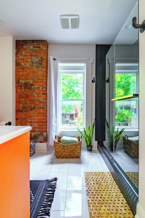 Sunlit modern bathroom with exposed red brick column, large window overlooking a leafy backyard, woven towel basket with folded towels, potted snake plant, glossy white floor tiles and glass shower with black herringbone tile accent.