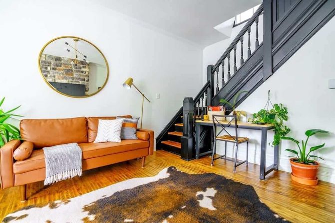 Bright modern living room with a tan leather sofa, patterned pillows and throw, round gold mirror, black wooden staircase, small desk and chair, potted plants, and cowhide rug on hardwood floors.