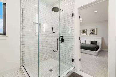 Bright modern ensuite with glass walk-in shower, white subway tile and matte-black rain and hand shower, opening into a contemporary master bedroom with gray bedding.